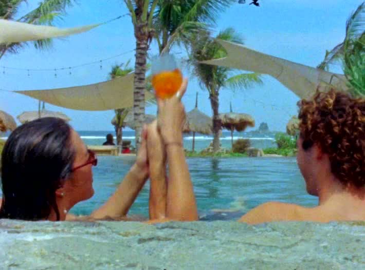Two people at the edge of an infinity pool hold an Aperol up between them, palms and shade sails over the water behind.