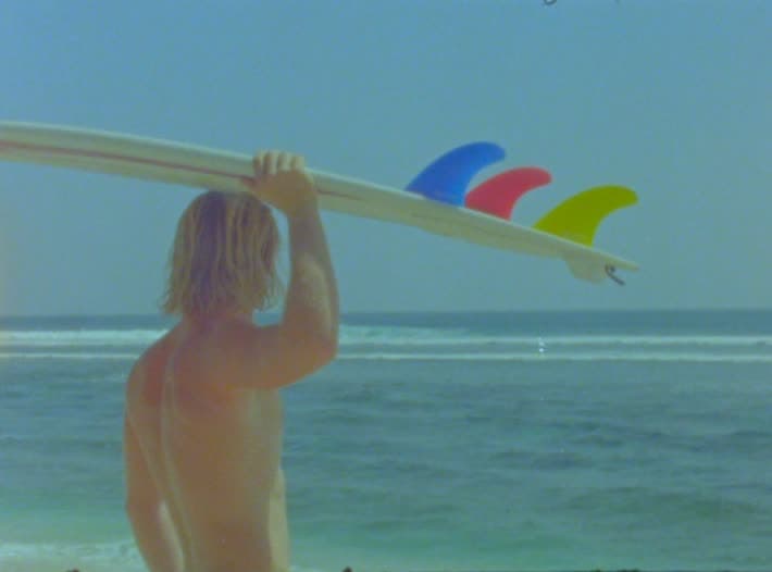 A surfer stands at the shore holding a white board overhead, its blue, red and yellow fins against a bleached sky, with a clean line-up breaking behind him.