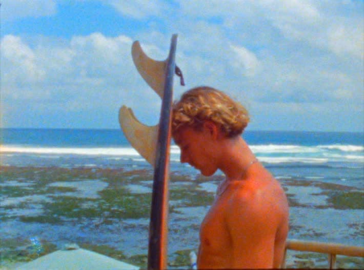 A surfer stands with his board held upright beside him, head down, looking at the reef and the line of white water behind.