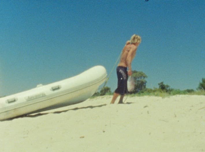 A figure drags an inflatable dinghy up a white sand bank by its bow line, scrub and deep blue sky behind.