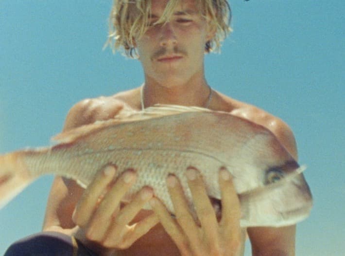 Close on a snapper held up in both hands, with the face of the spearfisher behind it against a bleached blue sky.