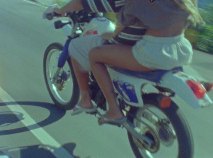 Two riders sharing a dirt bike down a sunlit road, shot low from alongside, their shadow running across the tarmac beside them.