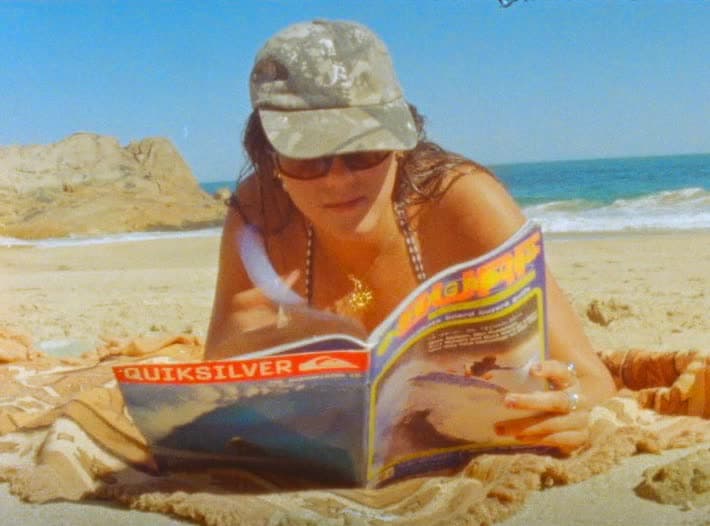 A woman lies on a towel in a cap and sunglasses, reading a surf magazine on the sand with the shore break behind her.