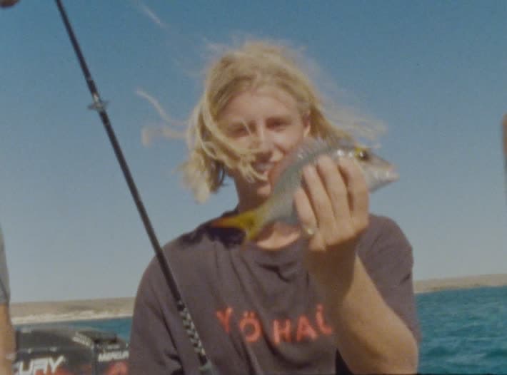 A young man on a boat holds a small striped fish up to camera, hair blown across his face, rod and outboard motor behind him.