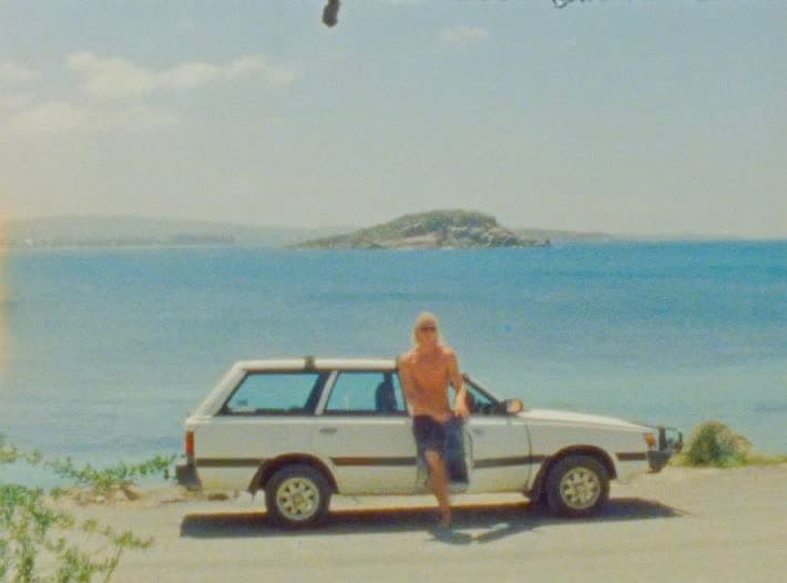 A man leans against a white station wagon parked on sand, an island sitting low on the water behind him.