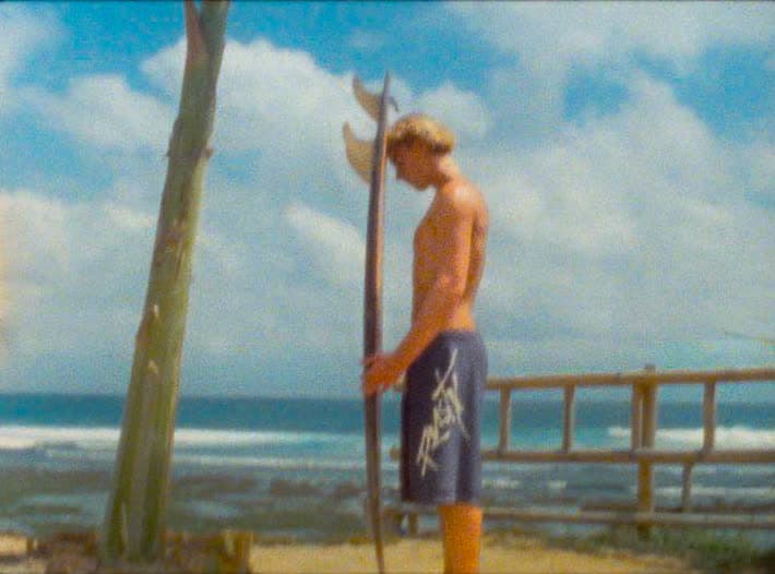 A surfer stands beneath a bare pole with a longboard held upright beside him, looking down at the shallow reef break behind.