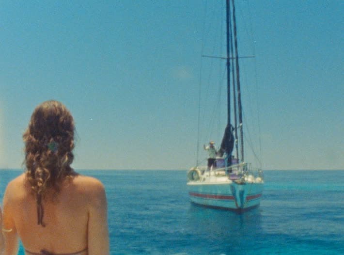 A woman with her back to camera watches a small yacht lying at anchor on flat blue water.