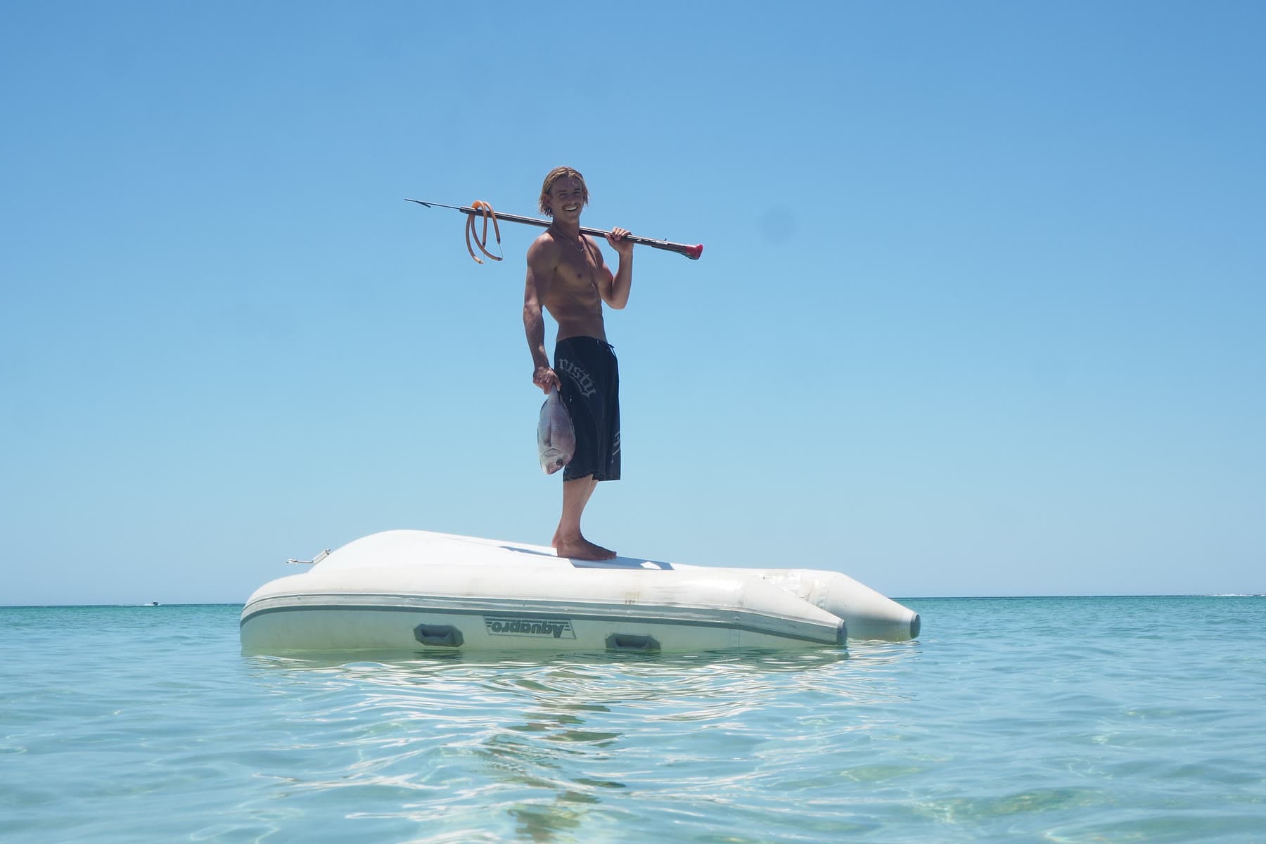 A spearfisher stands on a drifting inflatable dinghy in open water, speargun over one shoulder and a fish hanging from his hand.