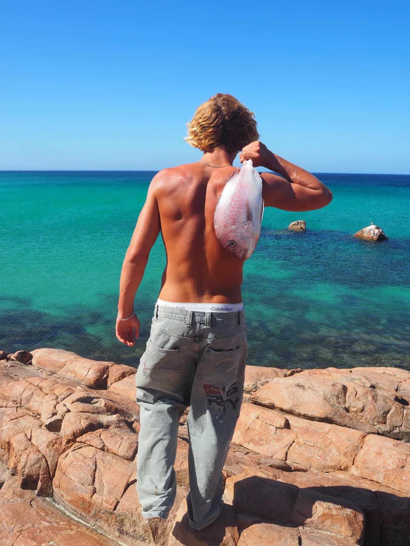 A spearfisher photographed from behind, a snapper slung over his bare shoulder against banded blue and green water.