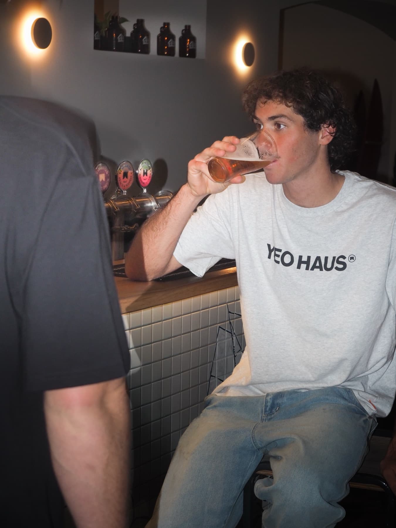 A drinker in a Yeo Haus shirt tips a glass of beer back at the bar, wall lights glowing behind him and growlers on the shelf above.