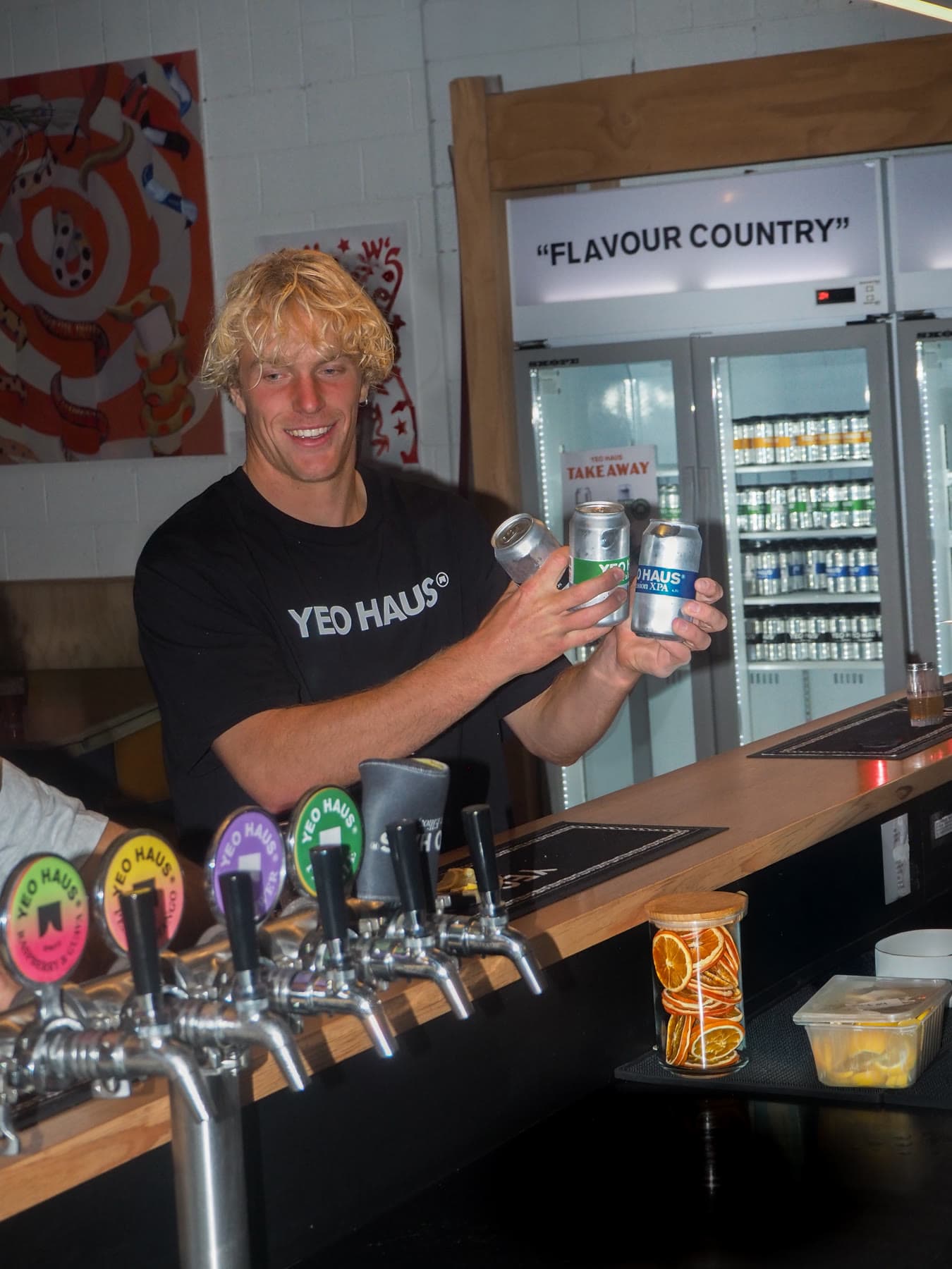 A bartender in a Yeo Haus shirt laughs behind the taps, holding three cans up over the bar with the cold room behind him.