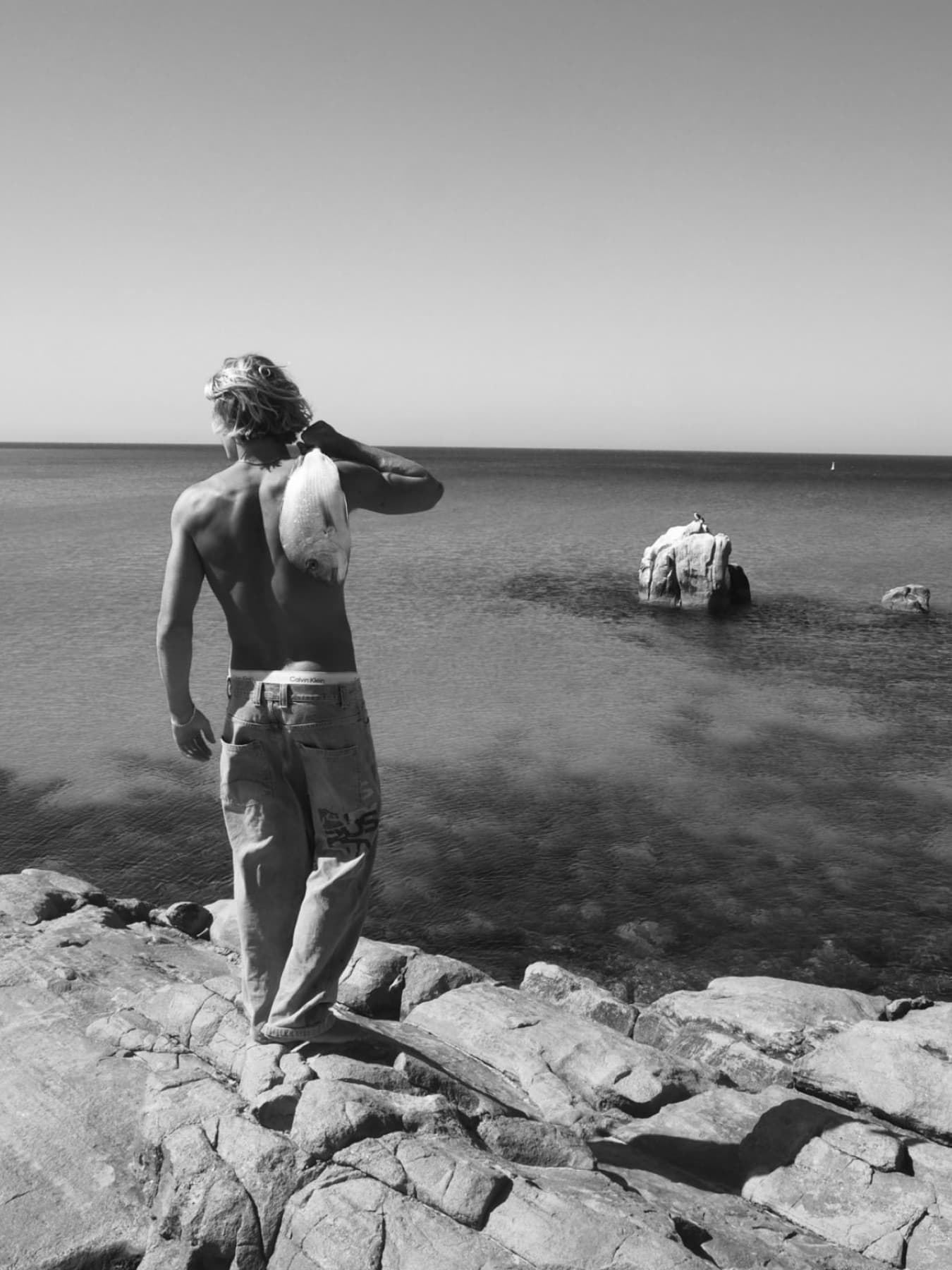 In black and white, a spearfisher stands with his back to the camera on the rocks, a fish over his shoulder, looking out at a rock in the flat water.
