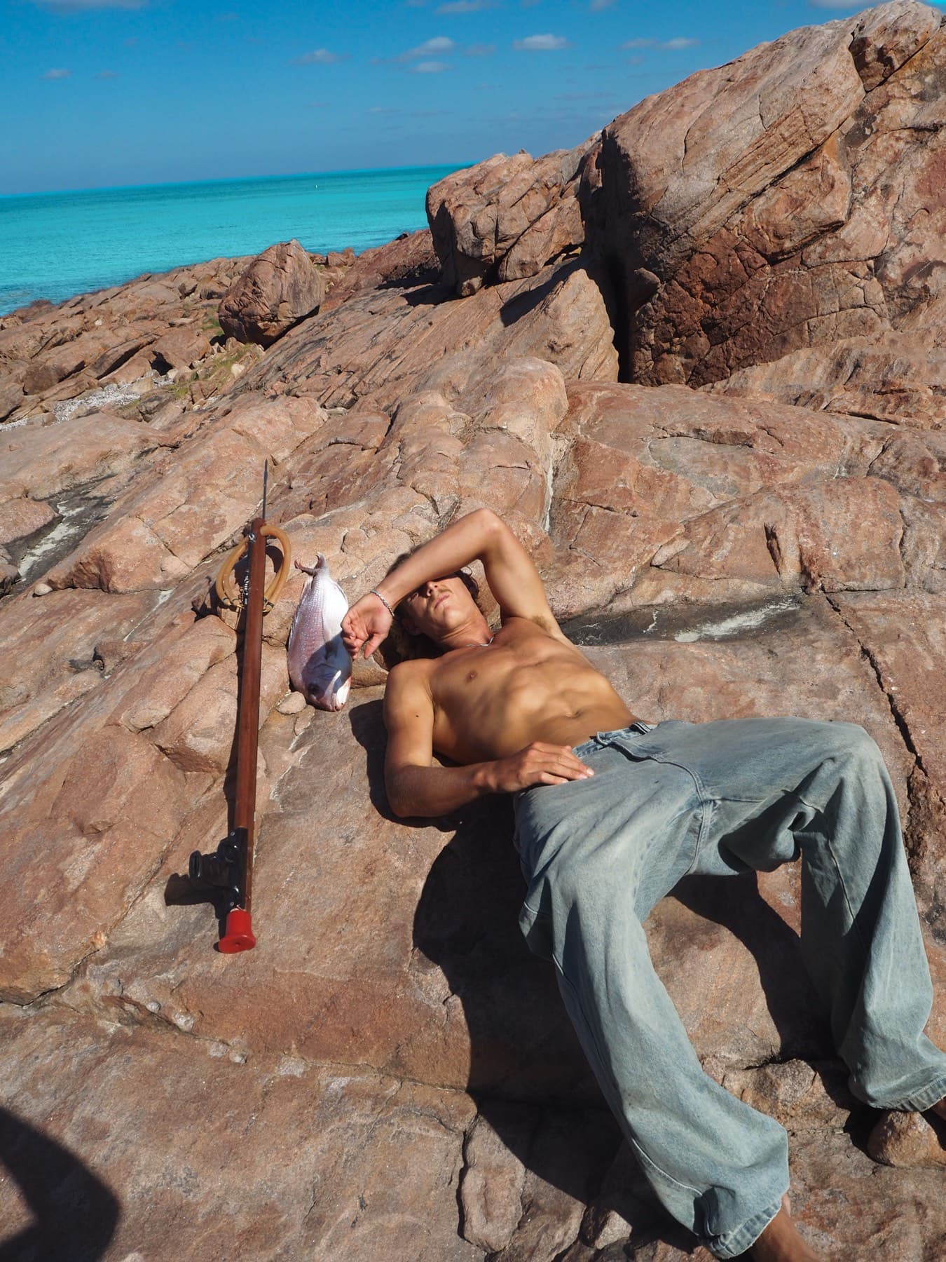 A spearfisher lies back on sunlit granite, one arm shading his eyes, speargun and catch resting on the rock beside him.