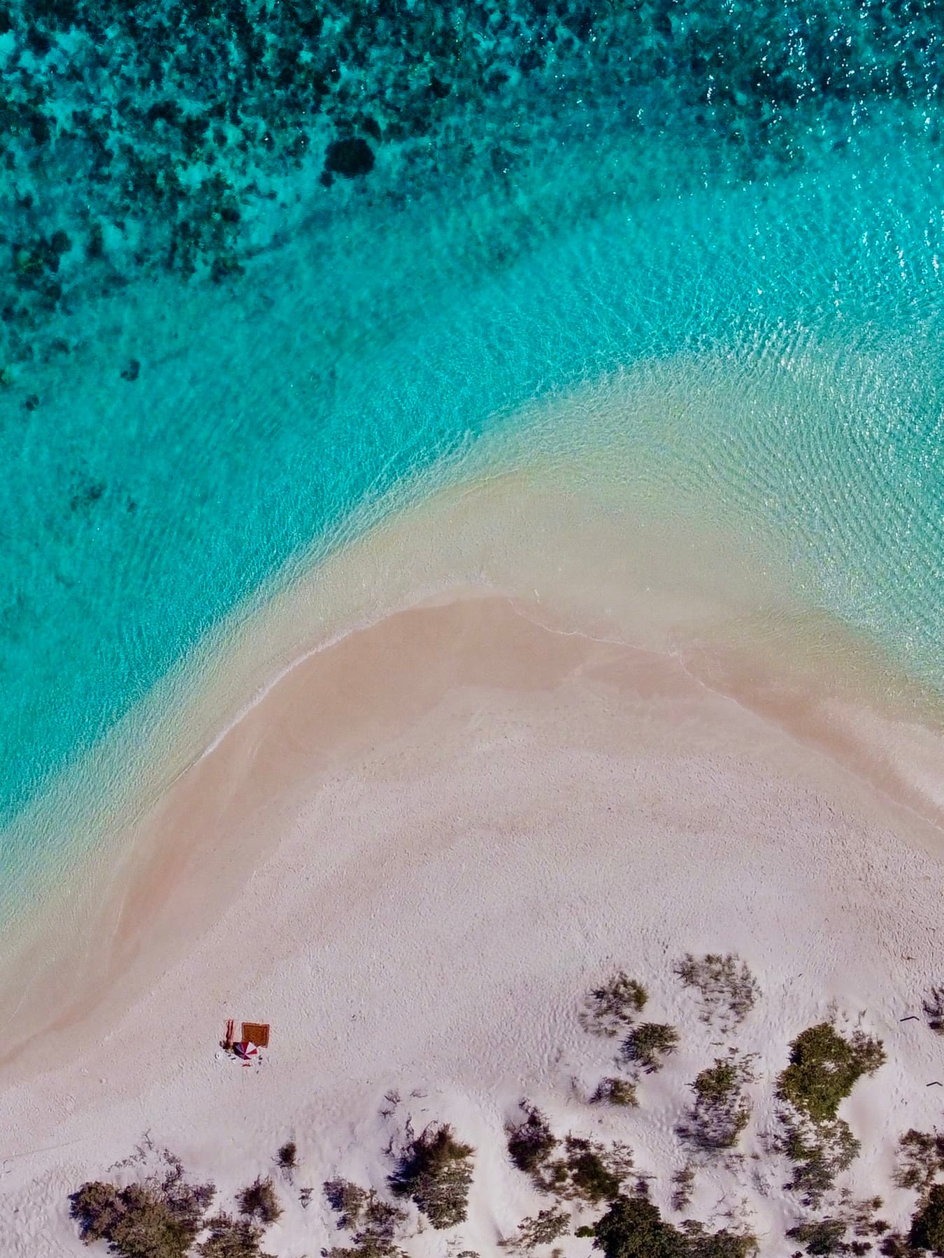 Straight down from above: a curved white sandbar dividing pale shallows from dark turquoise reef, two people on a towel small against the sand.