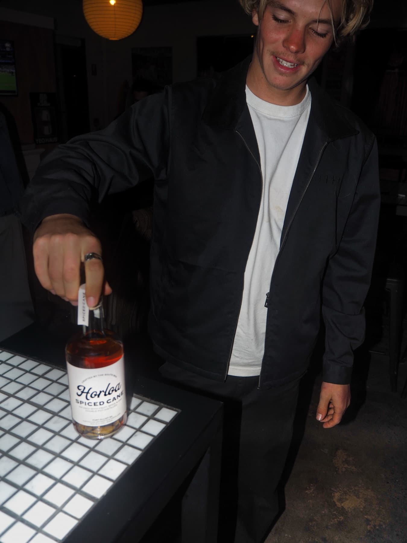 A man in a black jacket grins as he sets a bottle of Horloa Spiced Cane rum down on a white tiled table in a dark bar.