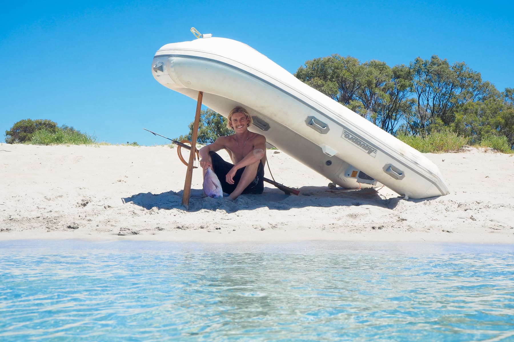A spearfisher sits in the shade of an inflatable dinghy propped on an oar, his catch in the sand beside him and clear shallows in the foreground.