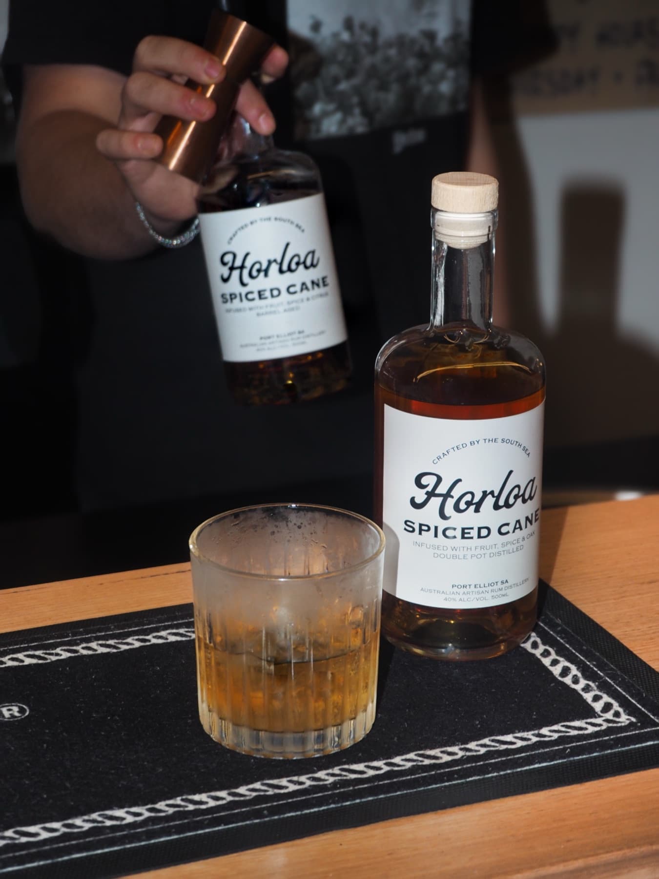 A bottle of Horloa Spiced Cane rum stands beside a tumbler of rum on ice on a bar mat, a bartender tipping a second bottle in the dark behind them.