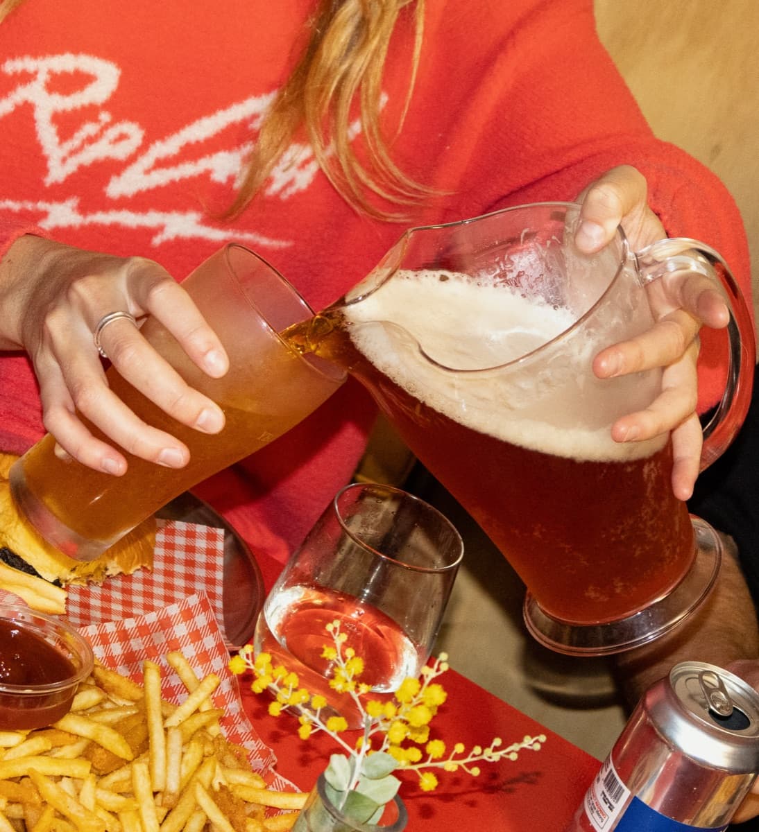 A woman in a red jumper pours beer from a full jug into a tilted glass, chips and a can of Yeo Haus on the table below.