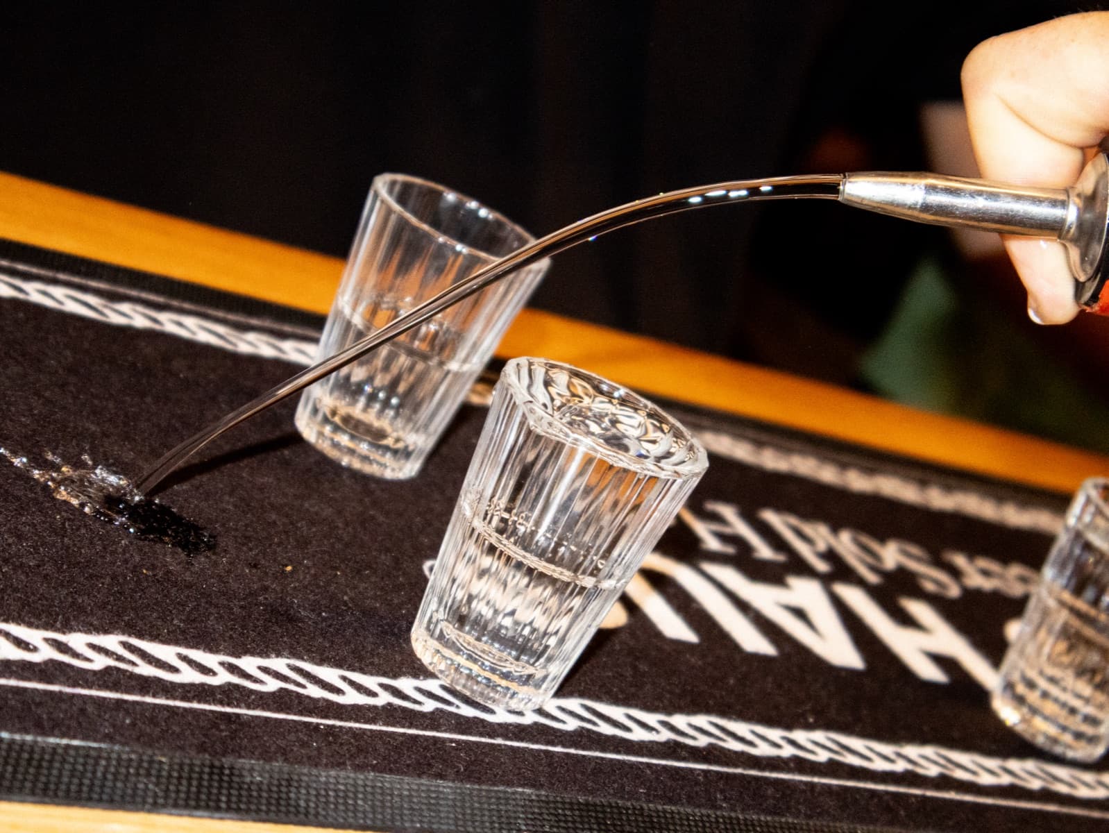 Close on two shot glasses on a Yeo Haus bar mat, a long metal pourer filling the second from above.