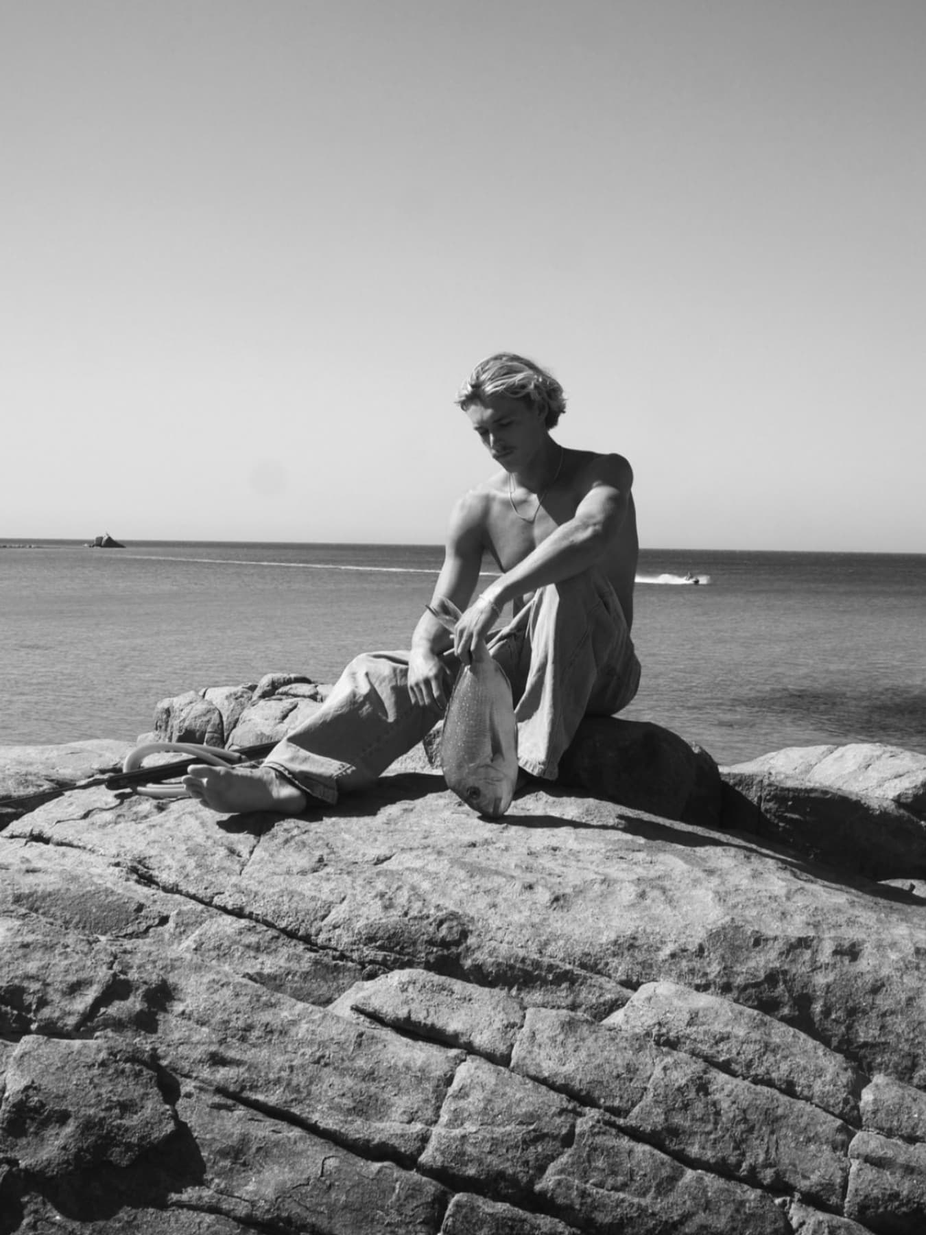 In black and white, a spearfisher sits barefoot on a granite shelf holding a fish up by the gill, his speargun on the rock beside him.