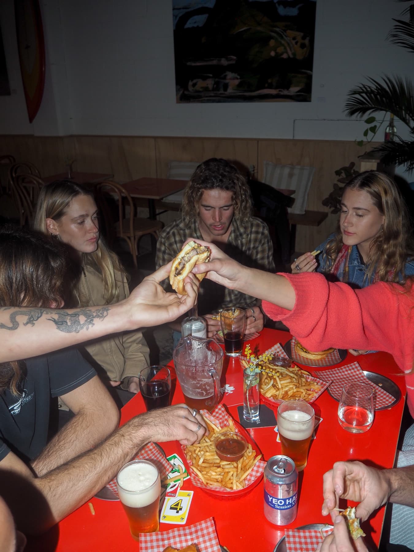 Friends crowd a red table passing a burger between them, baskets of chips, pints and a Yeo Haus can spread across it.