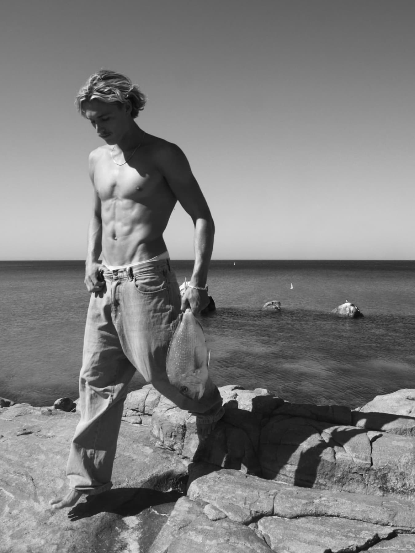 In black and white, a spearfisher walks down a rock shelf with a fish hanging from one hand, flat sea and small rocks behind him.
